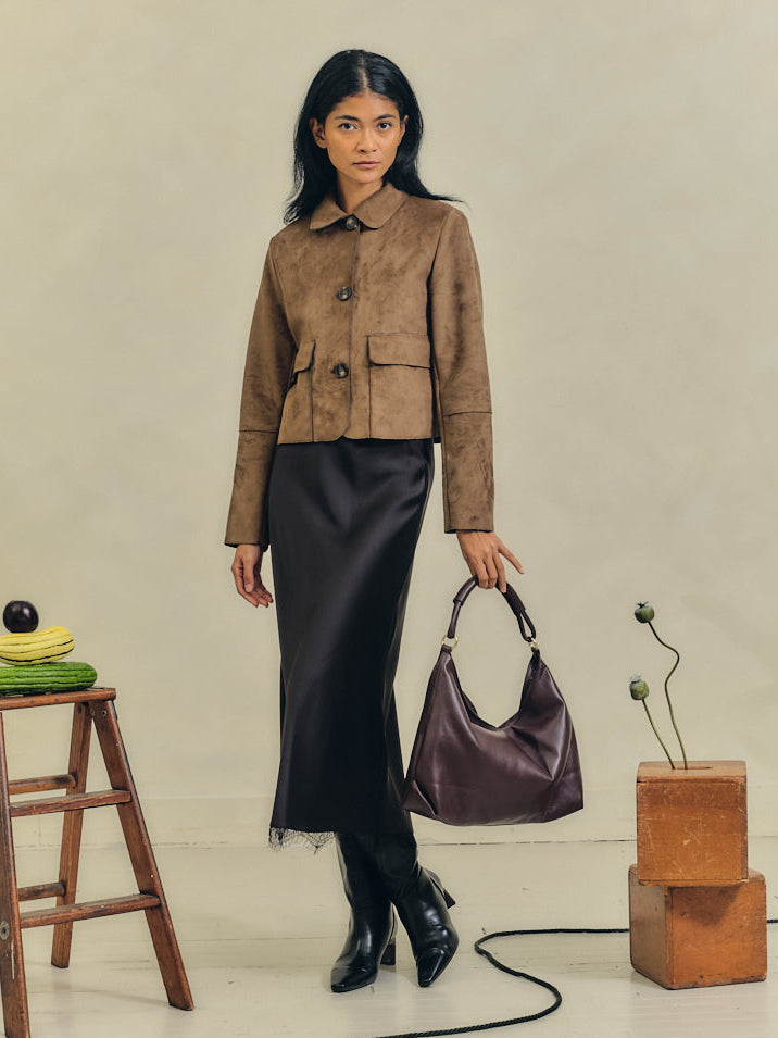 Woman in a brown jacket and black skirt standing in a minimalistic room with furniture and objects.